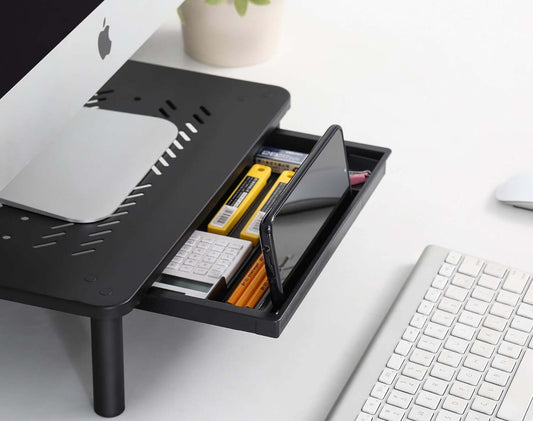 Adjustable metal monitor stand in black with an open drawer, holding office supplies and a phone, placed on a white desk with a keyboard.